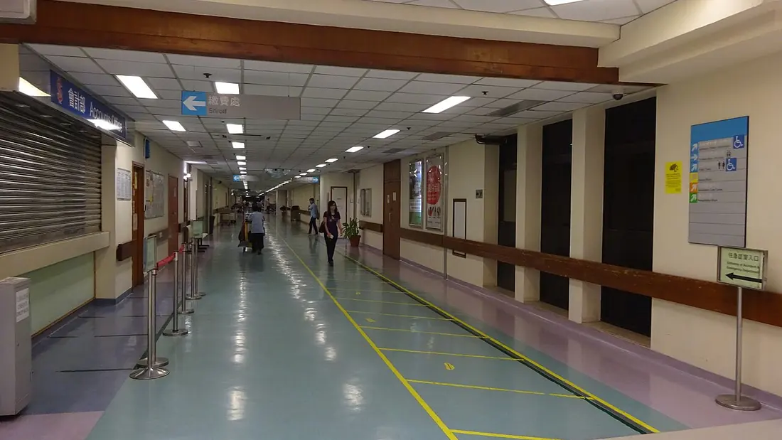 A wide hospital corridor with overhead signs and queue posts