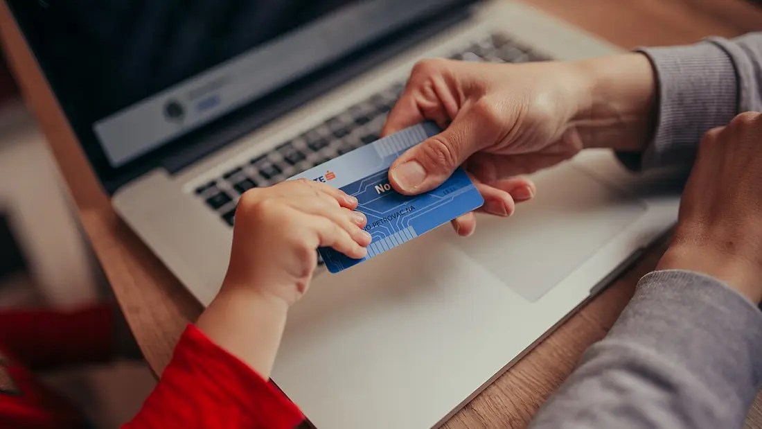 A child’s hand and an adult’s hand holding a bank card over a laptop
