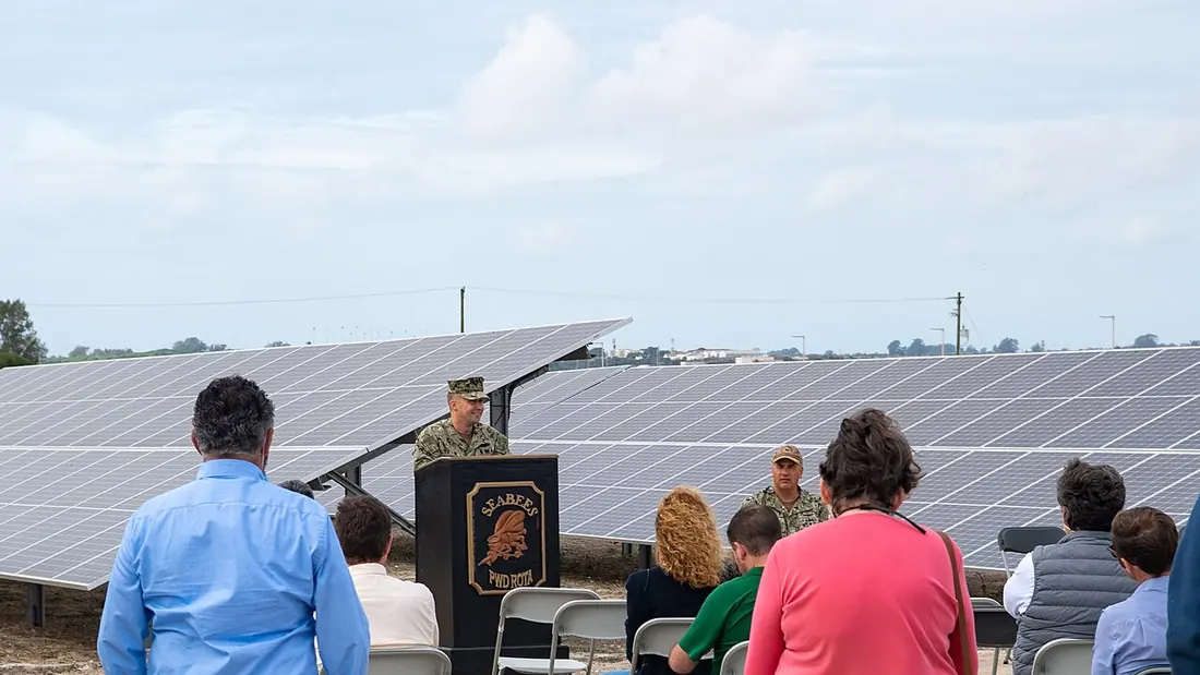 A speaker at a lectern in front of long rows of photovoltaic panels